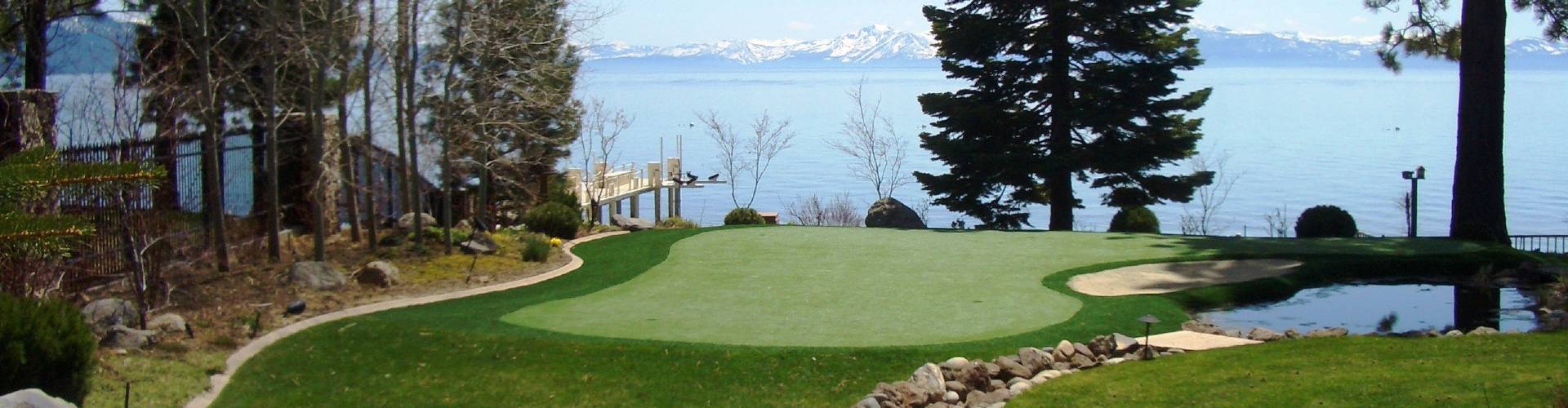 Outdoor putting green with a sand bunker and rock border installed at a lakefront estate with a private dock, pine trees, and snow-capped mountains in the background